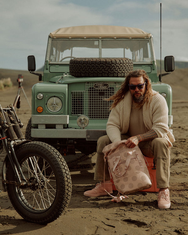 Jason Momoa wearing the Dirty Pink Chukkas while sitting on a cooler in front of a Land Rover.