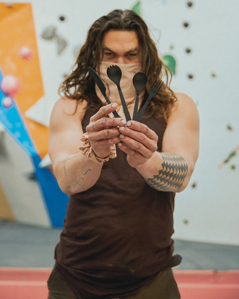 Jason Momoa holding a set of black wolf cutlery in  front of his face while in a climbing gym.